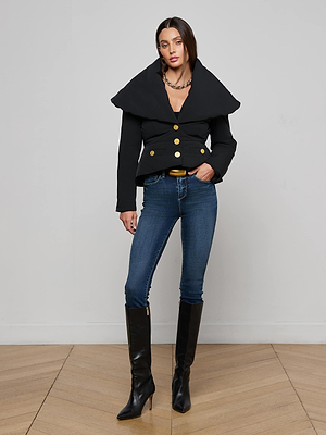 A woman with straight brown hair poses with one hand in her pocket against a white wall, wearing the black Pippa High Collar Jacket, blue skinny jeans, and knee-high black heeled boots.