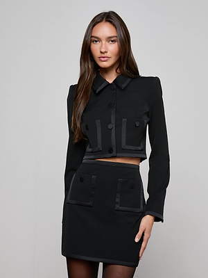 A woman with long brown hair models the Blythe Cropped Jacket, a stylish black blazer with buttons and large front pockets, paired with a matching skirt against a plain light gray background.