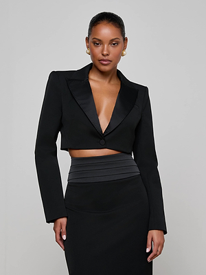 A woman stands against a plain background wearing the L'AGENCE Daxton Cropped Blazer—black with satin lapels—over a high-waisted black skirt. She accessorizes with pearl earrings, has sleek pulled-back hair, and exudes confidence.
