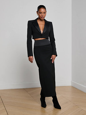 A woman stands indoors on a wooden floor, wearing the L’AGENCE Daxton Cropped Blazer with a satin lapel and deep V-neck, styled with a matching long black skirt and ankle boots for an effortlessly chic eveningwear look.