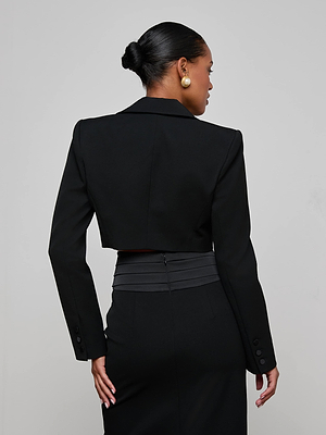 A woman with her hair in a neat bun models the L’AGENCE Daxton Cropped Blazer, featuring a satin lapel. Turned away, she reveals the back of her evening look and large gold stud earrings, set against a plain light gray background.