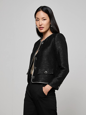 A woman with long dark hair models the L’AGENCE Zell Sequined Tweed Jacket, featuring decorative buttons and trim, over a light top and black pants against a plain background, with one hand in her pocket.