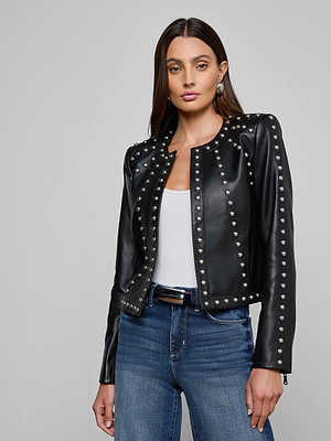 A woman confidently poses against a plain gray background, wearing the Fia Studded Leather Jacket with silver studs over a white top and blue jeans.
