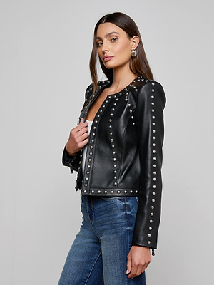 A woman with long brown hair models the Fia Studded Leather Jacket, blue jeans, and silver hoop earrings. She stands against a plain gray background, looking confidently at the camera.
