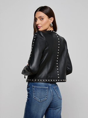 A woman with long brown hair poses in the Fia Studded Leather Jacket, featuring silver studs, paired with blue jeans, looking over her shoulder against a plain gray background.