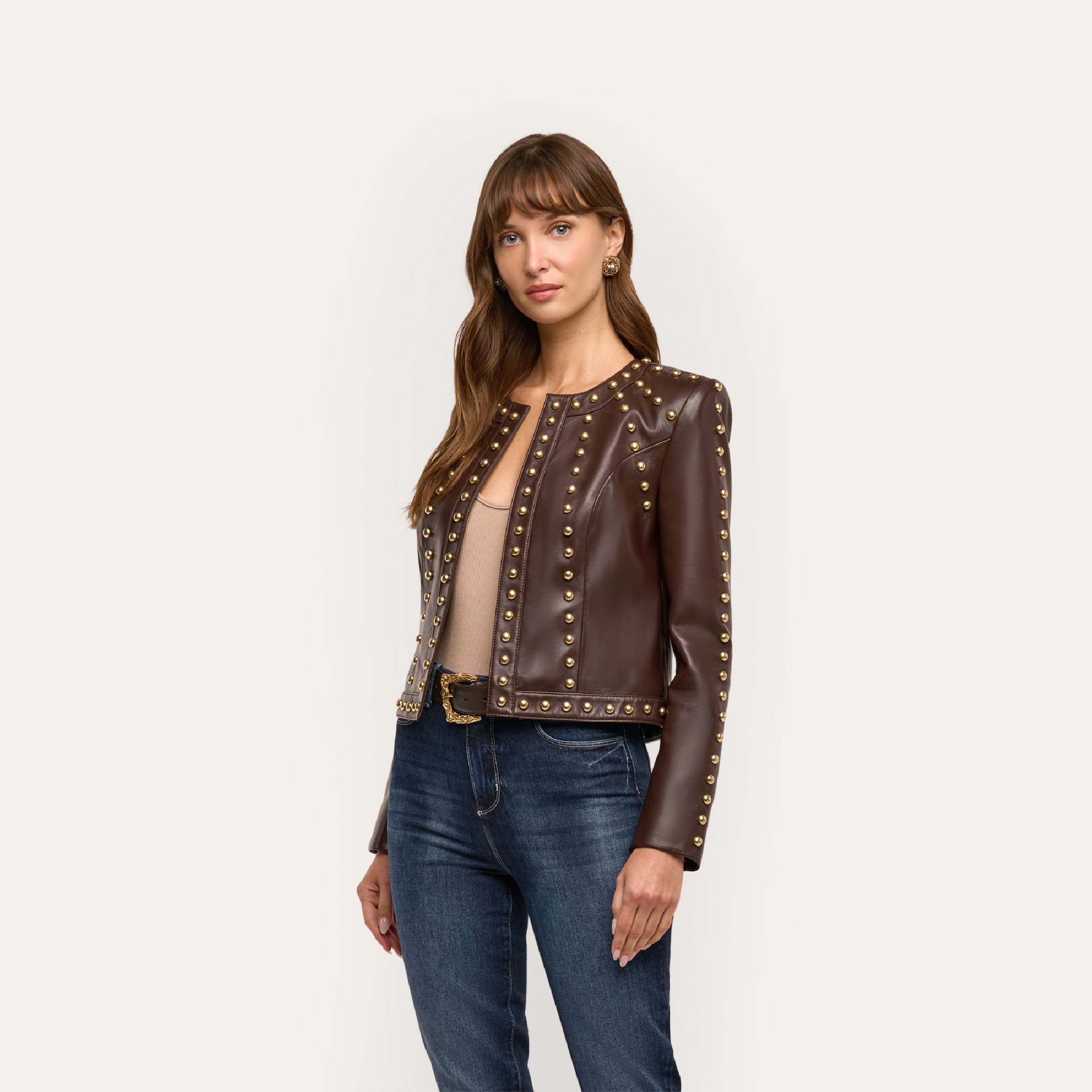 A woman wears a chocolate brown lambskin Fia Leather Jacket with gold studs along the collarless neckline, front opening, and hem, paired with blue jeans.