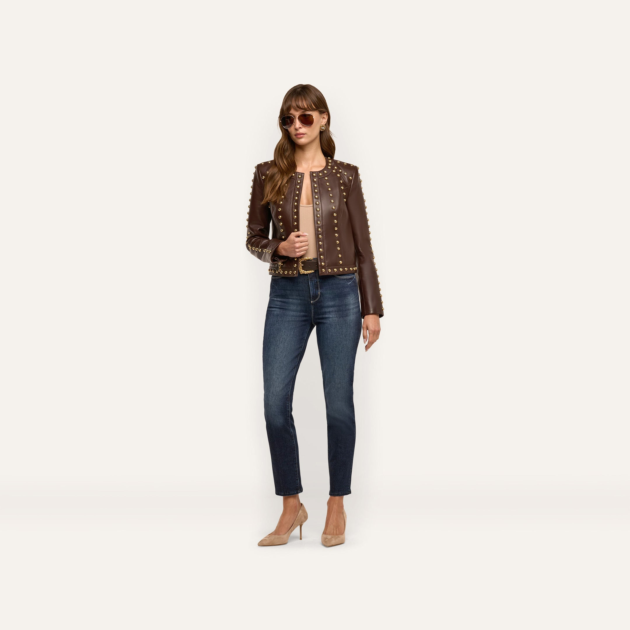 A woman wears a chocolate brown, studded Fia Leather Jacket paired with dark blue jeans and tan high-heeled pumps.
