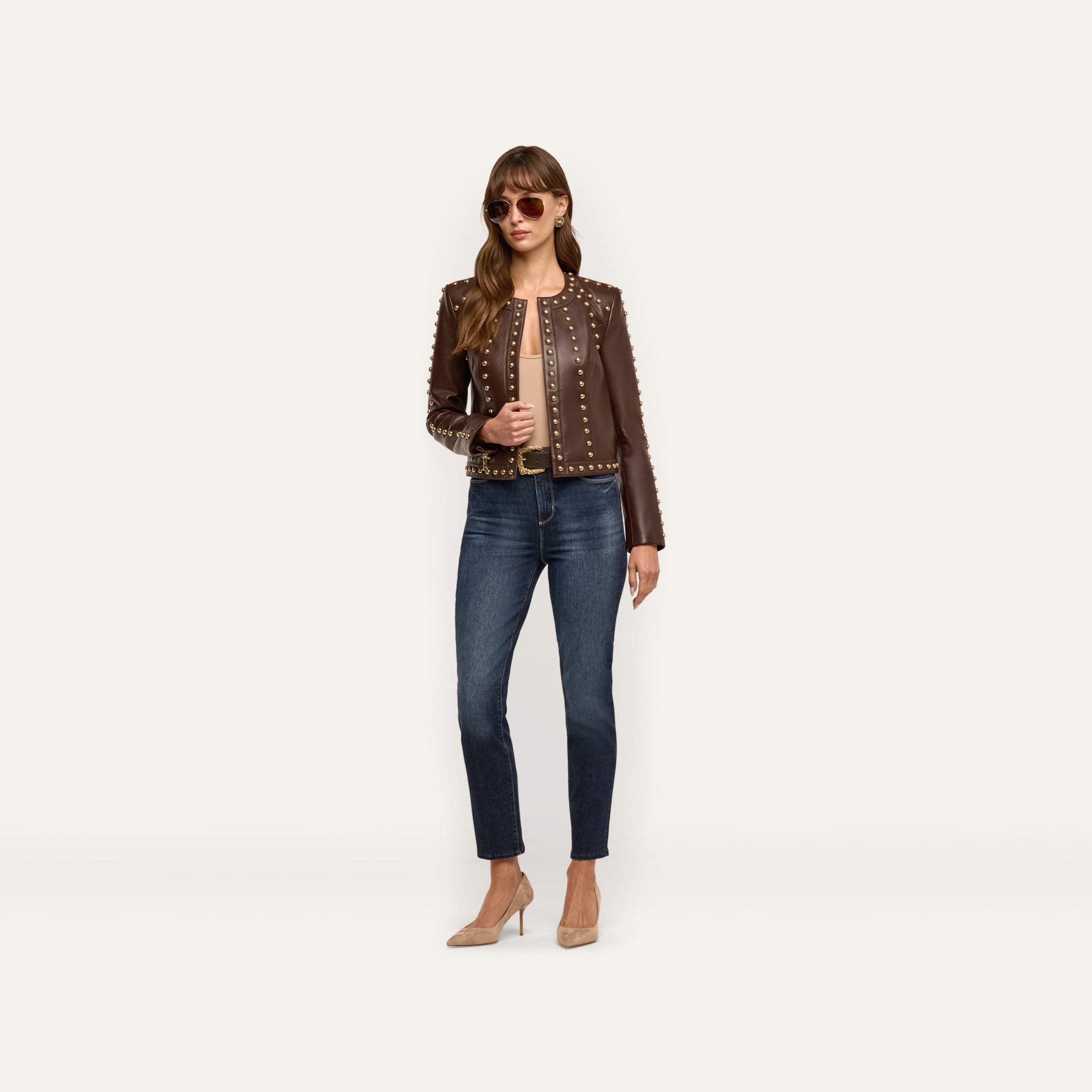 A woman wears a chocolate brown, studded Fia Leather Jacket paired with dark blue jeans and tan high-heeled pumps.