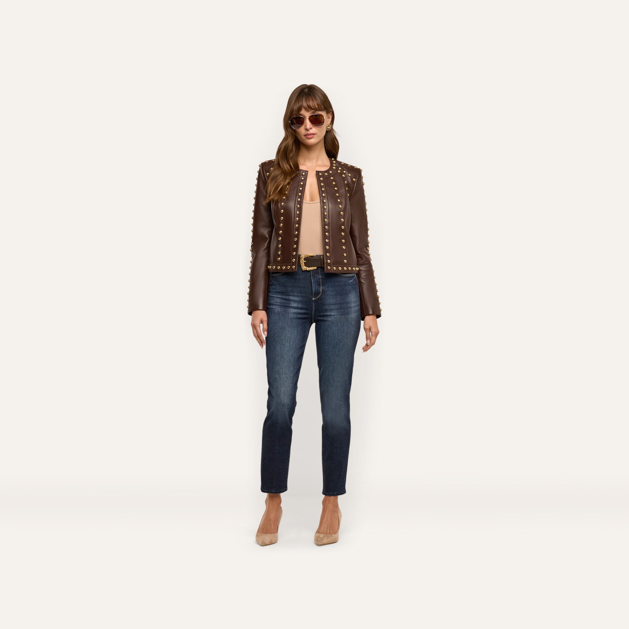 A model wearing a chocolate brown lambskin Fia Leather Jacket with gold stud detailing, a tan top, blue jeans, and tan heels.