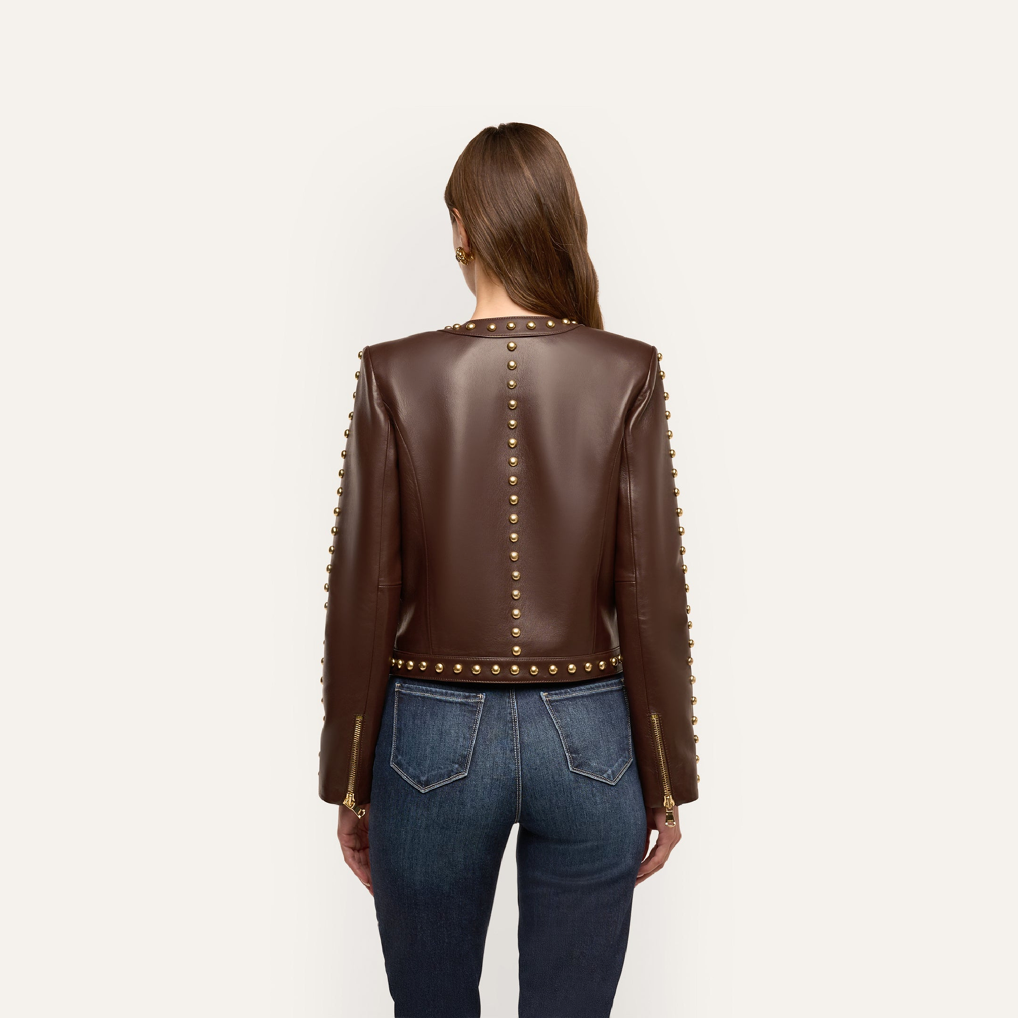 Back view of the chocolate brown Fia Leather Jacket with gold embellishments along the neckline, center back, and sleeves, worn with dark blue jeans.