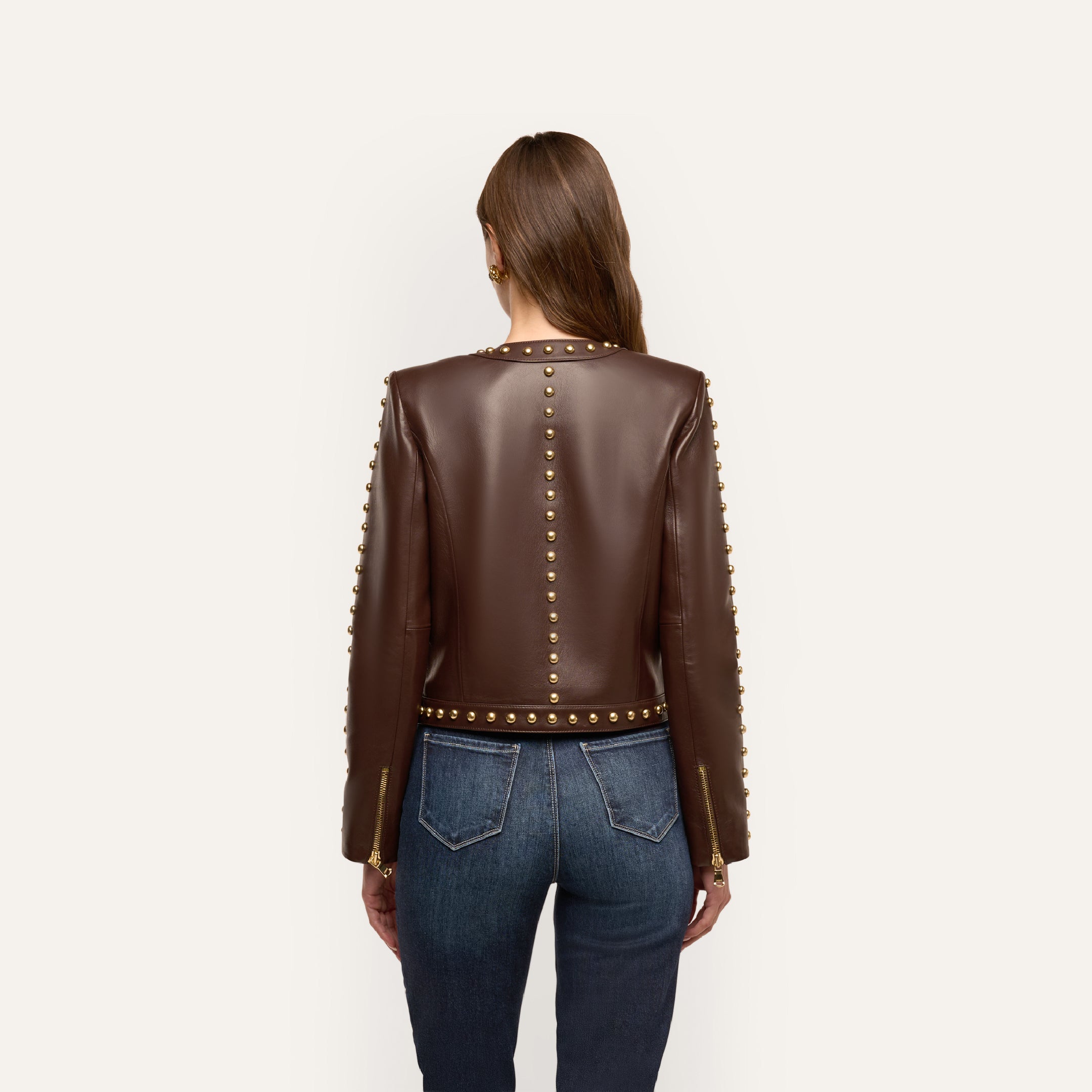 Back view of the chocolate brown Fia Leather Jacket with gold embellishments along the neckline, center back, and sleeves, worn with dark blue jeans.