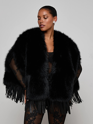 A model poses against a light gray background wearing the L'AGENCE Nathalia Faux Fur Cape, a dramatic black fringe-trimmed style, over a black mesh outfit. Her hair is slicked back and she accessorizes with stud earrings.