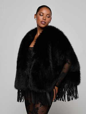 A woman poses against a plain light gray background, wearing the L'AGENCE Nathalia Faux Fur Cape with fringe details over a sheer black lace outfit.