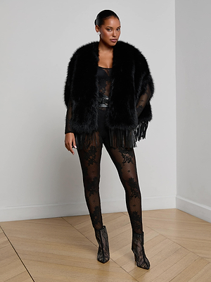 A woman stands in a corner of a room wearing a black sheer lace bodysuit, black heels, and the L'AGENCE Nathalia Faux Fur Cape. Her hair is pulled back as she poses confidently on the wooden floor.
