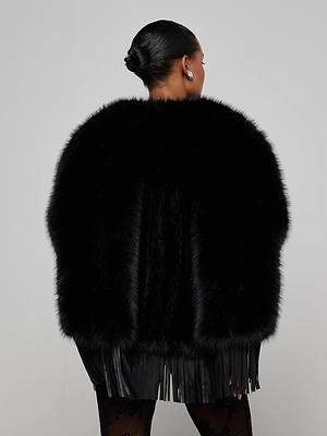 With their hair in a sleek bun, a person faces away from the camera, wearing the L'AGENCE Nathalia Faux Fur Cape featuring long black fringe at the hem, set against a plain light gray background.