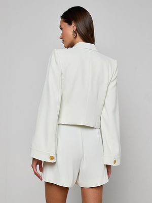 A woman with straight brown hair faces away, wearing the Nixie Cardigan Jacket—an open-front white blazer with gold buttons and matching high-waisted shorts—crafted from fluid suit fabric, set against a plain light background.