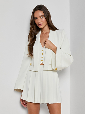 A woman with long brown hair models the Nixie Cardigan Jacket—an open-front blazer with wide bell sleeves in fluid suit fabric—paired with a white vest, pleated skirt, and gold buttons, standing by a white wall.