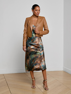 Indoors, a woman wears the Dulcie Leather Blazer in tan with whipstitch details over a shiny, multicolored feather-print slip dress. She accessorizes with gold heels, hoop earrings, and has her hair pulled back sleekly.