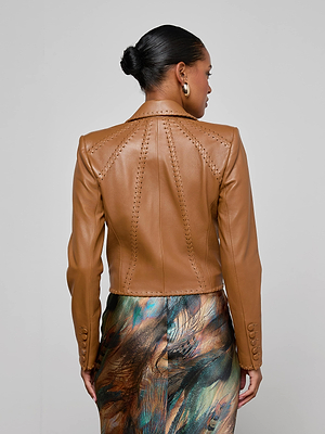 A woman with her hair in a sleek bun models the Dulcie Leather Blazer, a tan jacket with whipstitch detailing, paired with a multicolored abstract skirt. She stands facing away from the camera against a plain background.