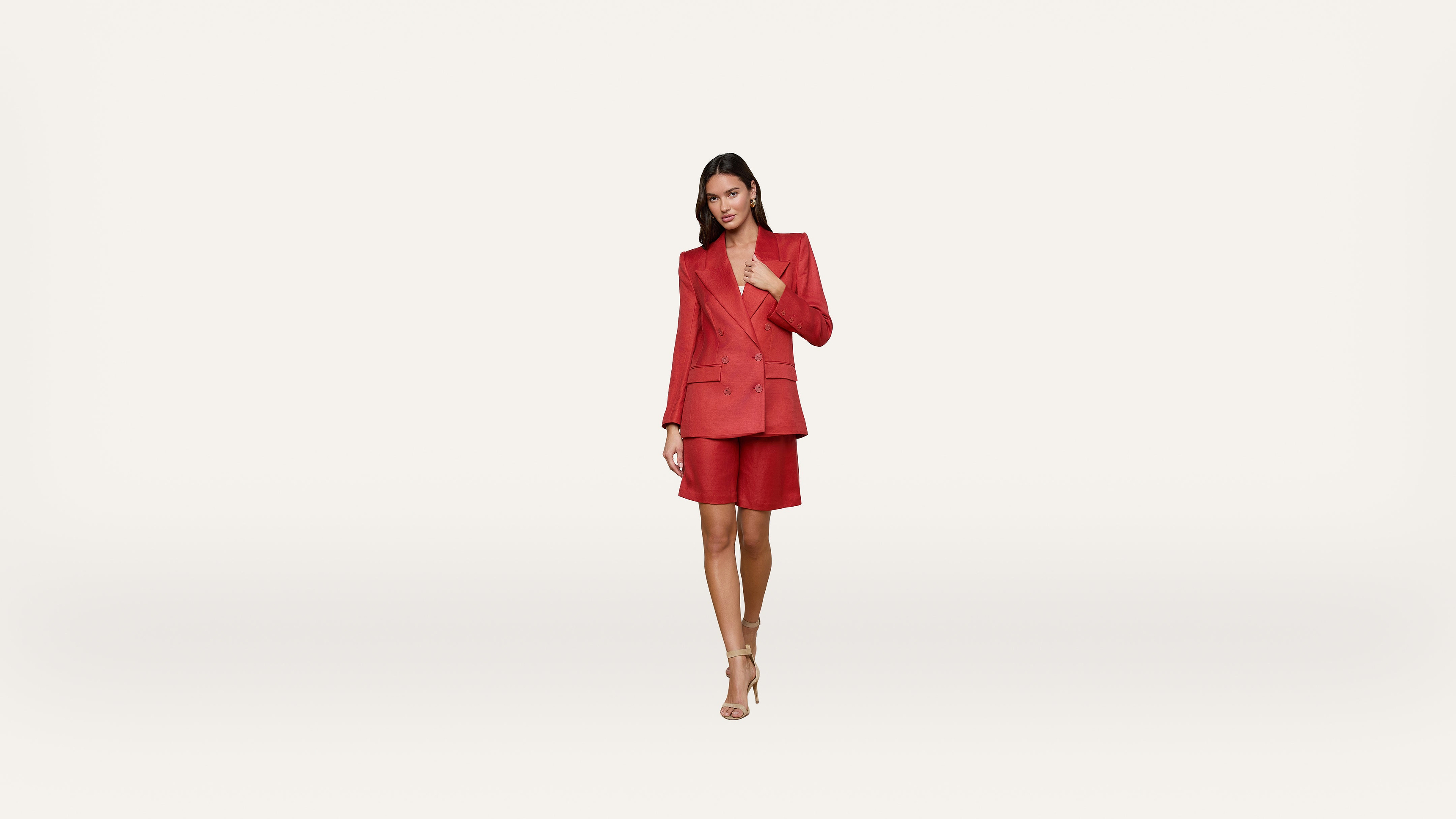 A woman stands against a plain white background, wearing a red blazer and matching shorts with nude heels, looking confidently at the camera.