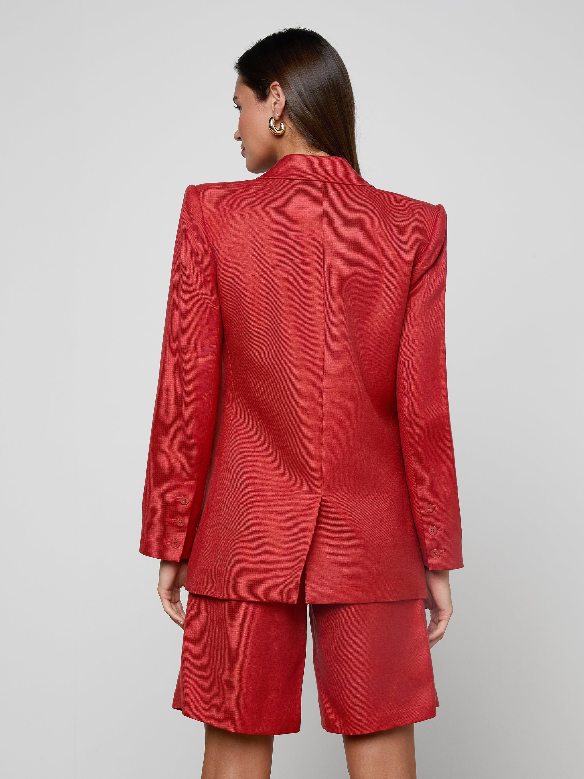 A woman with long brown hair is seen from behind against a plain light background, wearing the Cambrie Oversized Blazer in red paired with matching knee-length shorts.