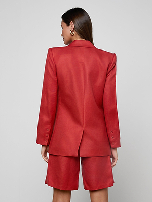 A woman with straight brown hair stands with her back to the camera, wearing the Cambrie Oversized Blazer in red and matching shorts, styled with gold hoop earrings against a plain, light background.