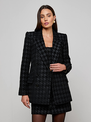 A woman with long dark hair wears the Cambrie Oversized Houndstooth Blazer and matching skirt, paired with gold earrings. She stands against a plain light gray background, looking confidently at the camera.