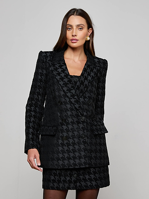 A woman with long brown hair wears the Cambrie Oversized Houndstooth Blazer and a matching skirt, standing against a plain light-gray background. She accessorizes with gold earrings and has a neutral expression.