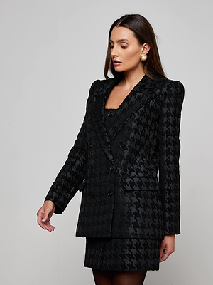 A woman with long brown hair and gold earrings models the Cambrie Oversized Houndstooth Blazer and matching skirt against a plain light gray background, looking slightly down and to the side.