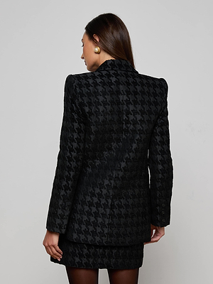 A woman with straight brown hair and gold earrings models the Cambrie Oversized Houndstooth Blazer with a matching skirt, standing facing away from the camera against a plain light background.