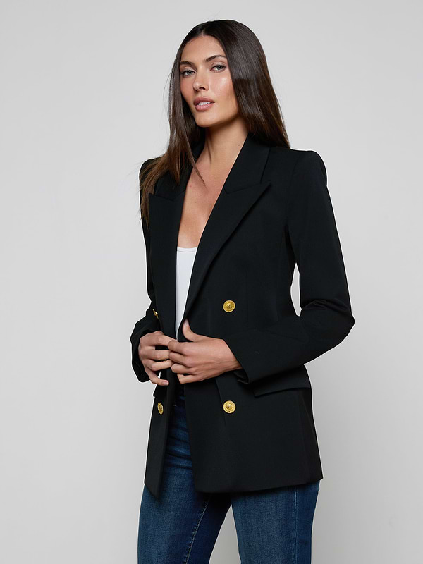 A woman with long brown hair wears the Cambrie Oversized Blazer—an oversized black blazer with gold buttons—paired with a white top and blue jeans, standing against a plain light background.