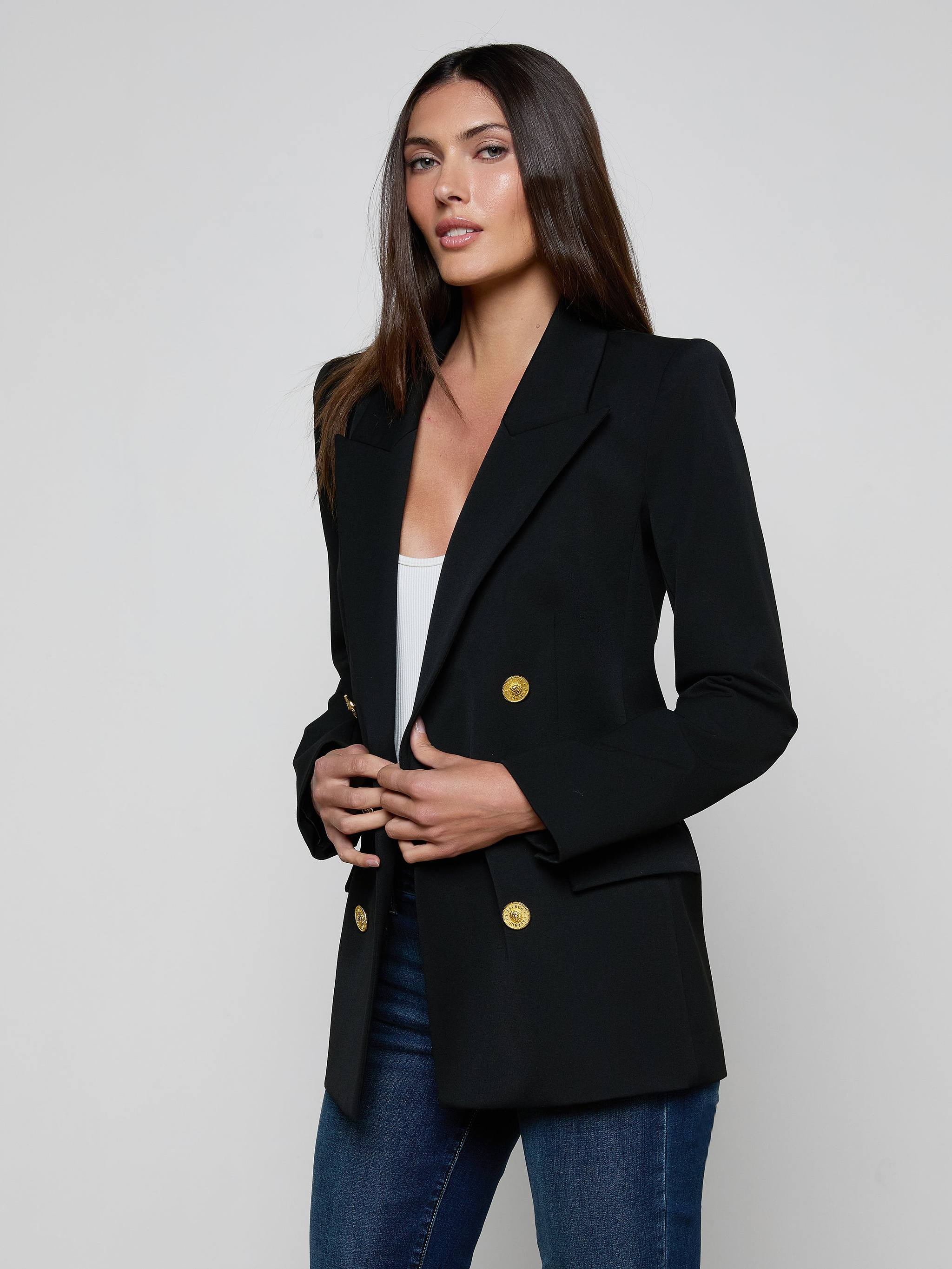 A woman with long brown hair wears the Cambrie Oversized Blazer—an oversized black blazer with gold buttons—paired with a white top and blue jeans, standing against a plain light background.