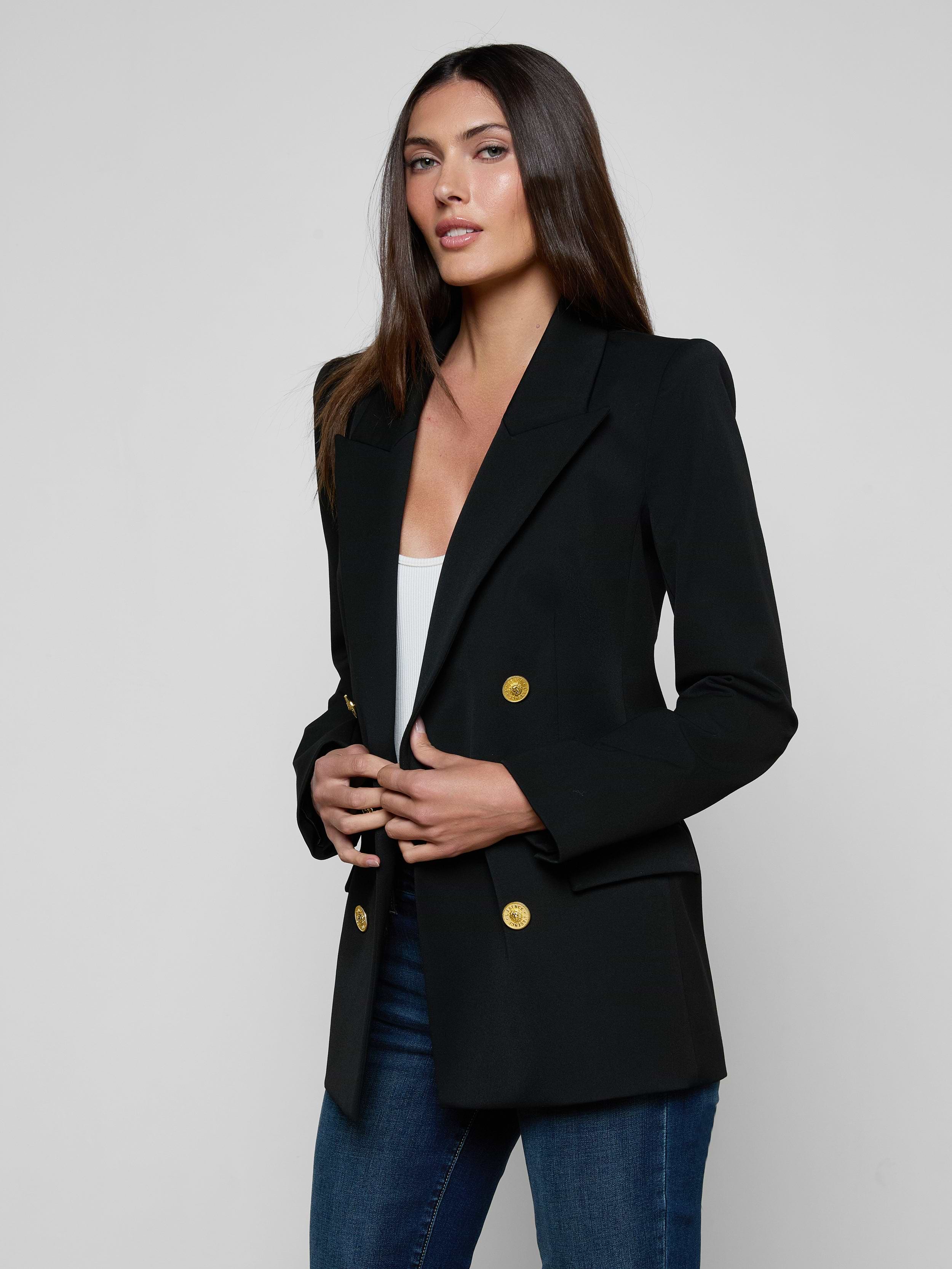 A woman with long brown hair wears the Cambrie Oversized Blazer—an oversized black blazer with gold buttons—paired with a white top and blue jeans, standing against a plain light background.