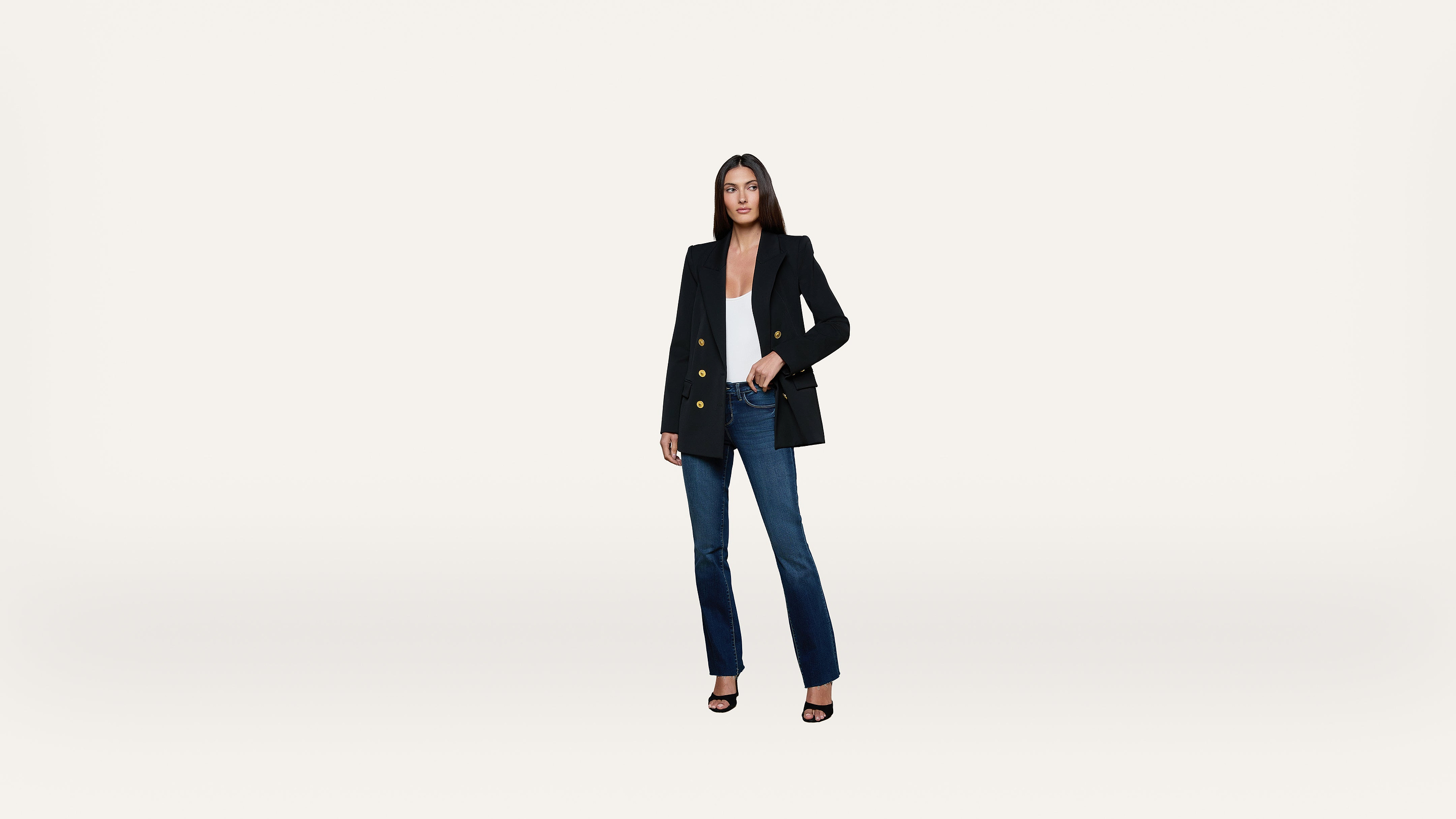 A woman stands against a plain white background wearing a black blazer, white top, blue jeans, and black heeled sandals, with one hand in her pocket.