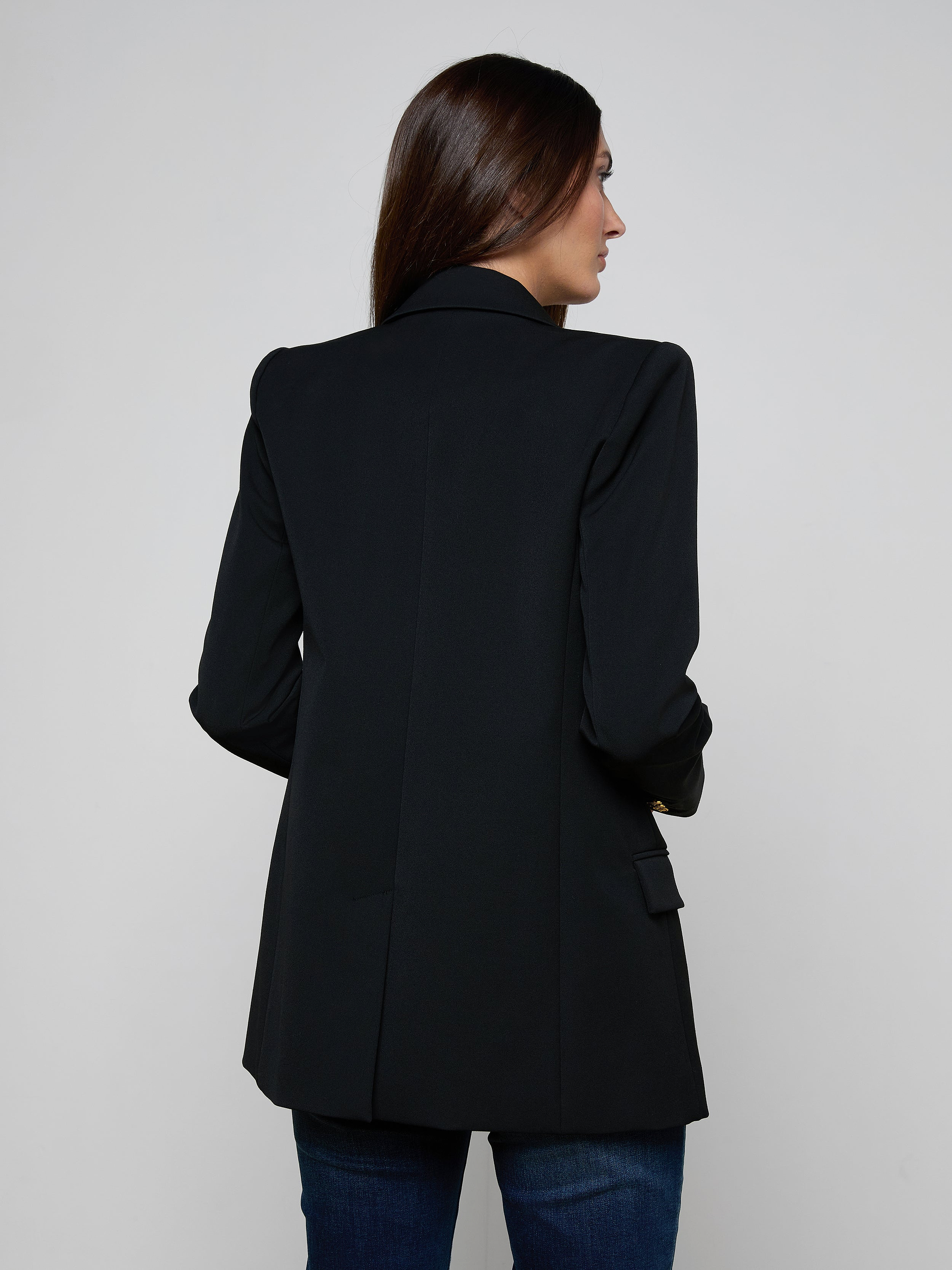 A woman with long brown hair stands facing away, showcasing the back of her outfit against a plain light background. She is wearing the Cambrie Oversized Blazer, crafted from stretch suit fabric, paired with blue jeans.