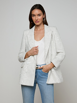 A woman stands against a light gray background wearing the Cambrie Oversized Sequin Blazer over a white top and blue jeans, one hand in her pocket and the other holding the blazer.