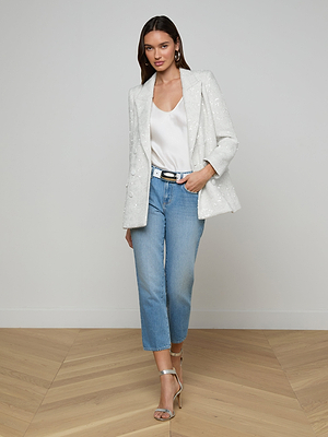 A woman with straight brown hair poses against a plain wall in the Cambrie Oversized Sequin Blazer, a white satin top, light blue cropped jeans, a black belt, and silver high-heeled sandals, one hand in her pocket.