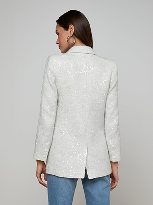 A woman with long dark hair wears the Cambrie Oversized Sequin Blazer with blue jeans and large hoop earrings, standing with her back to the camera against a plain gray background.
