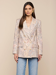 Une femme est tournée vers lavant, portant le blazer Cambrie Oversized Linen-Blend à gros boutons et un jean bleu clair, sur un fond beige uni.