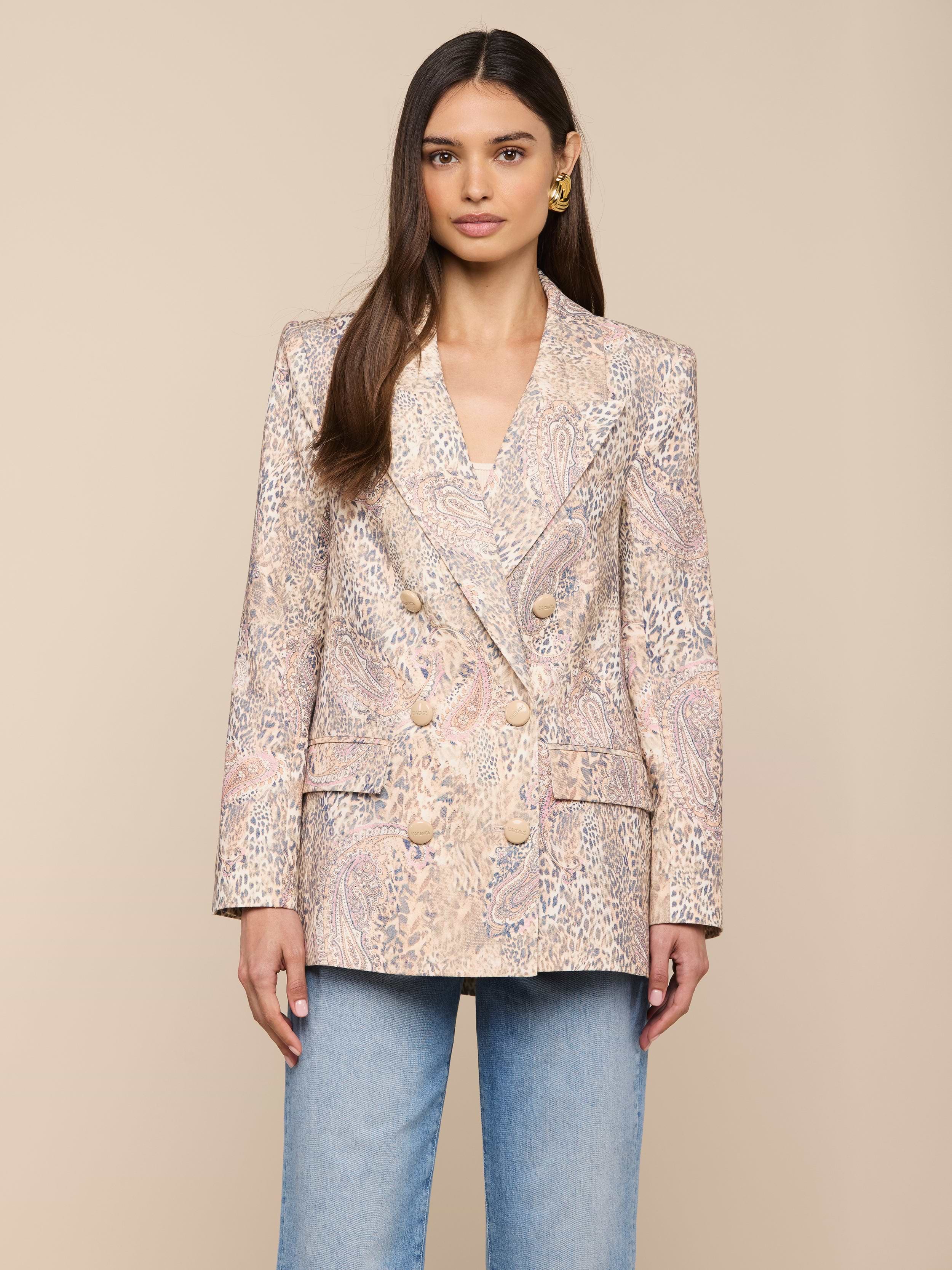 Une femme est tournée vers lavant, portant le blazer Cambrie Oversized Linen-Blend à gros boutons et un jean bleu clair, sur un fond beige uni.