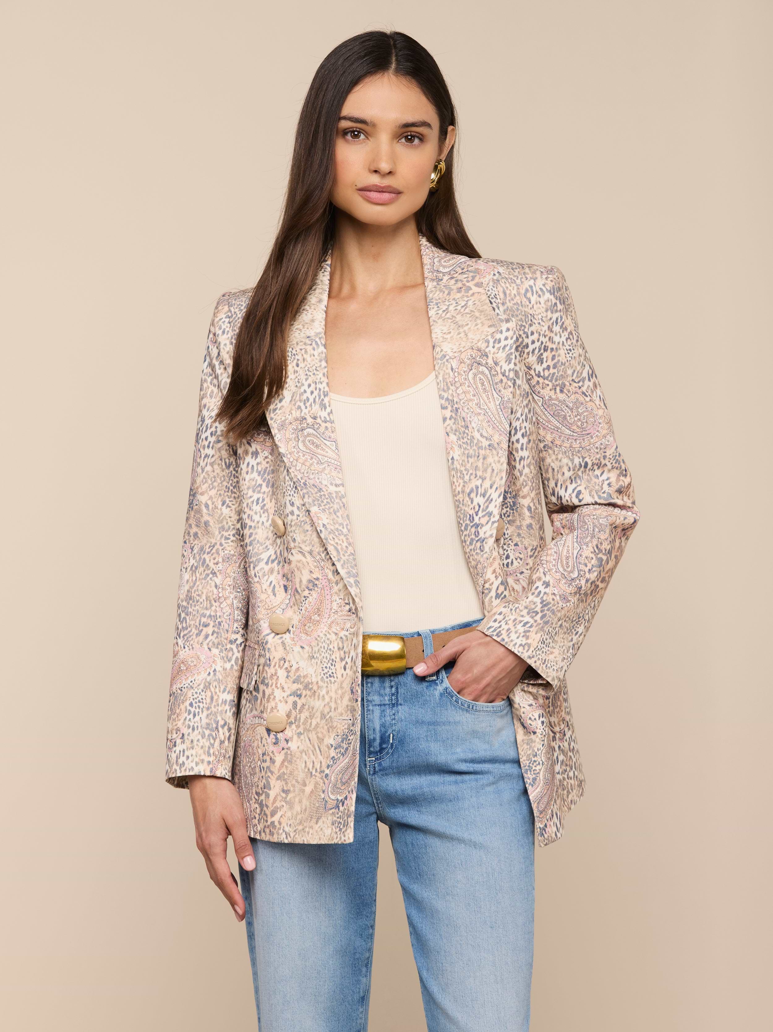 Une femme aux longs cheveux bruns porte le blazer Cambrie Oversized Linen-Blend sur un haut beige, un jean bleu clair et une ceinture dorée, sur un fond beige uni.