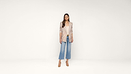 Woman standing against a plain white background, wearing a light patterned blazer, white top, wide-leg blue jeans, and tan heels. She has long brown hair and a neutral expression, with hands relaxed by her sides.