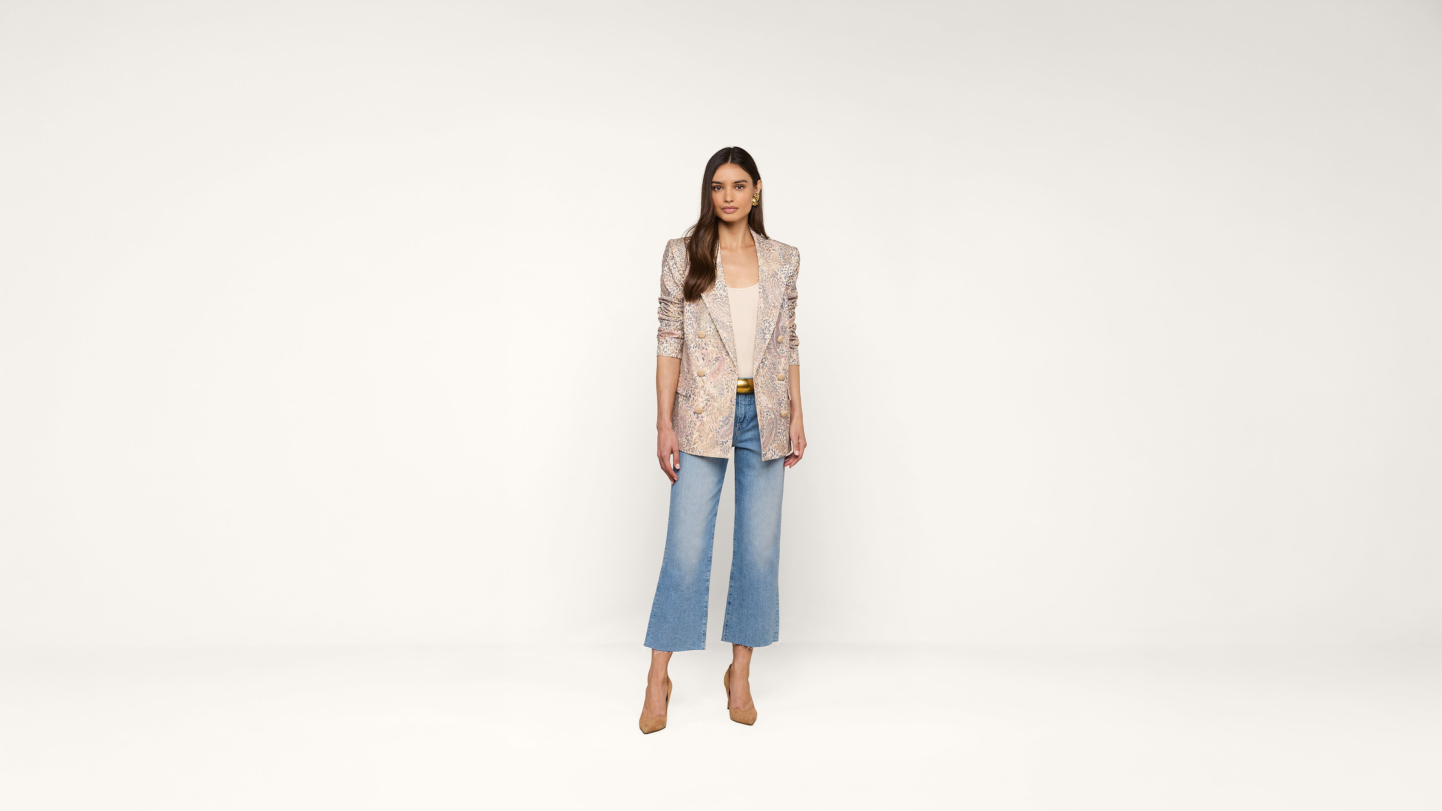 Woman standing against a plain white background, wearing a light patterned blazer, white top, wide-leg blue jeans, and tan heels. She has long brown hair and a neutral expression, with hands relaxed by her sides.
