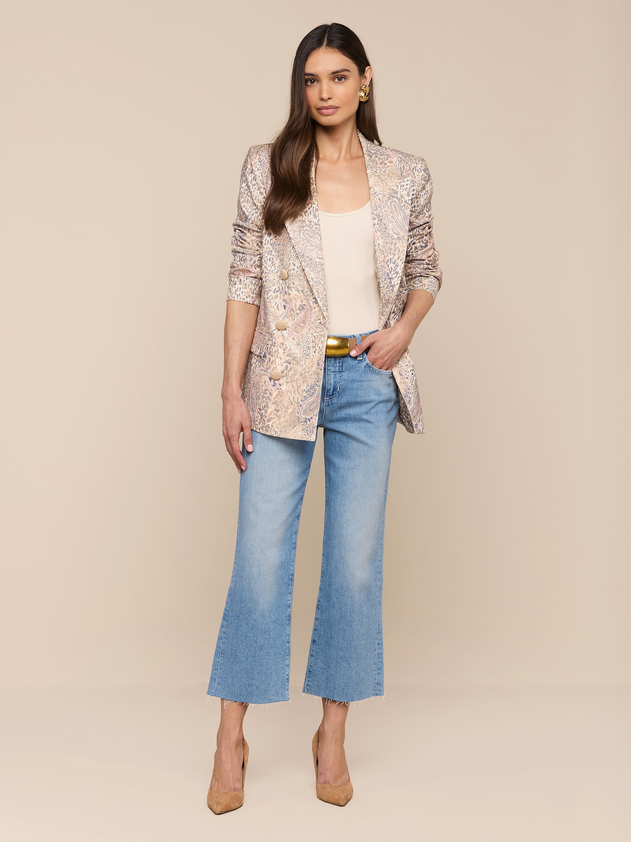 Une femme aux cheveux longs et raides et à lexpression neutre se tient sur un fond beige. Elle porte le blazer Cambrie Oversized Linen-Blend Blazer, un haut crème, un jean bleu, des talons à bouts pointus et des boucles doreilles dorées.