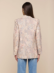 Une femme aux longs cheveux bruns est vue de dos, portant le blazer Cambrie Oversized Linen-Blend et un jean bleu clair, debout sur un fond beige clair uni.