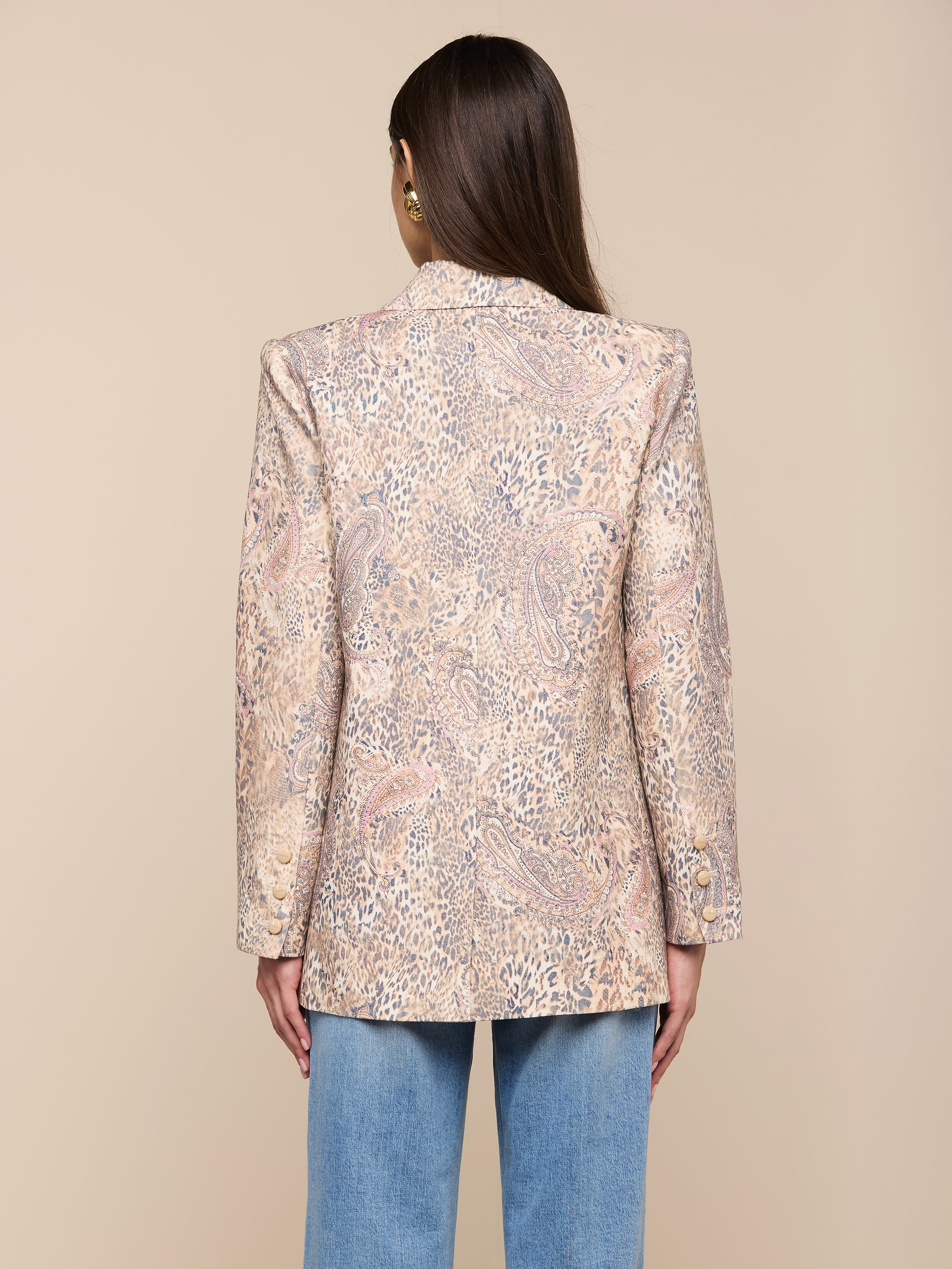 Une femme aux longs cheveux bruns est vue de dos, portant le blazer Cambrie Oversized Linen-Blend et un jean bleu clair, debout sur un fond beige clair uni.