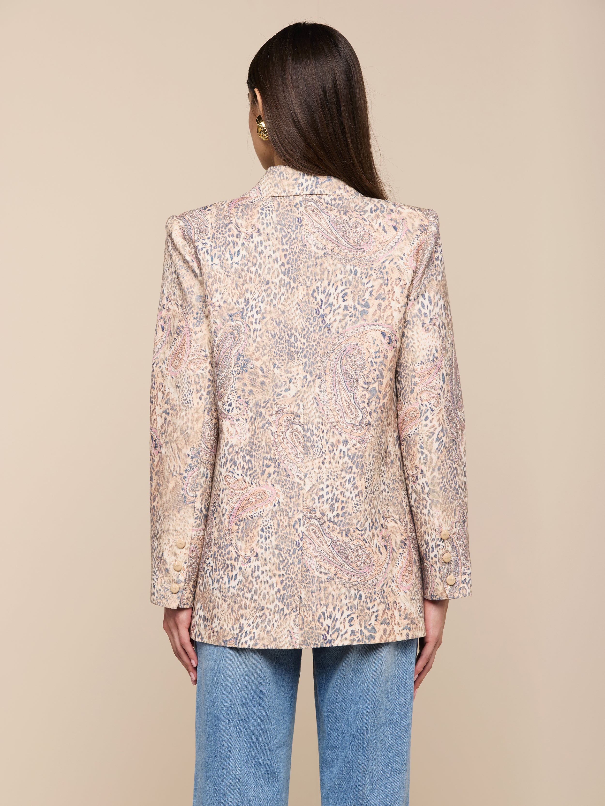 Une femme aux longs cheveux bruns est vue de dos, portant le blazer Cambrie Oversized Linen-Blend et un jean bleu clair, debout sur un fond beige clair uni.