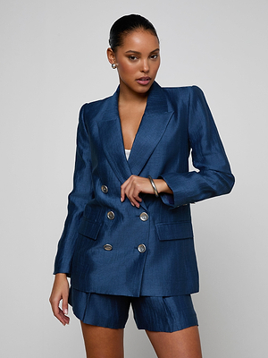 A woman poses against a plain background in the Cambrie Oversized Blazer—a dark blue piece with silver buttons—styled with matching shorts, her hair pulled back, and accessorized with earrings and a bracelet.