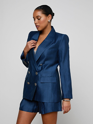 A woman wearing the Cambrie Oversized Blazer in dark blue with matching shorts stands against a plain background, gazing down and to the side while touching her lapel.