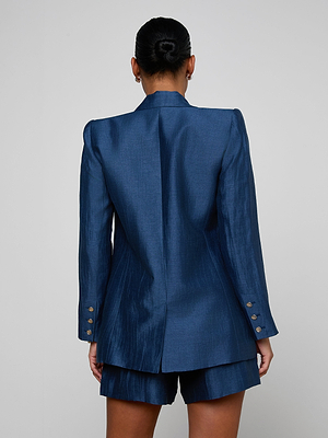 A person with dark hair in a neat bun is seen from behind, wearing the tailored Cambrie Oversized Blazer in shiny dark blue with gold buttons and matching shorts, set against a plain light background.